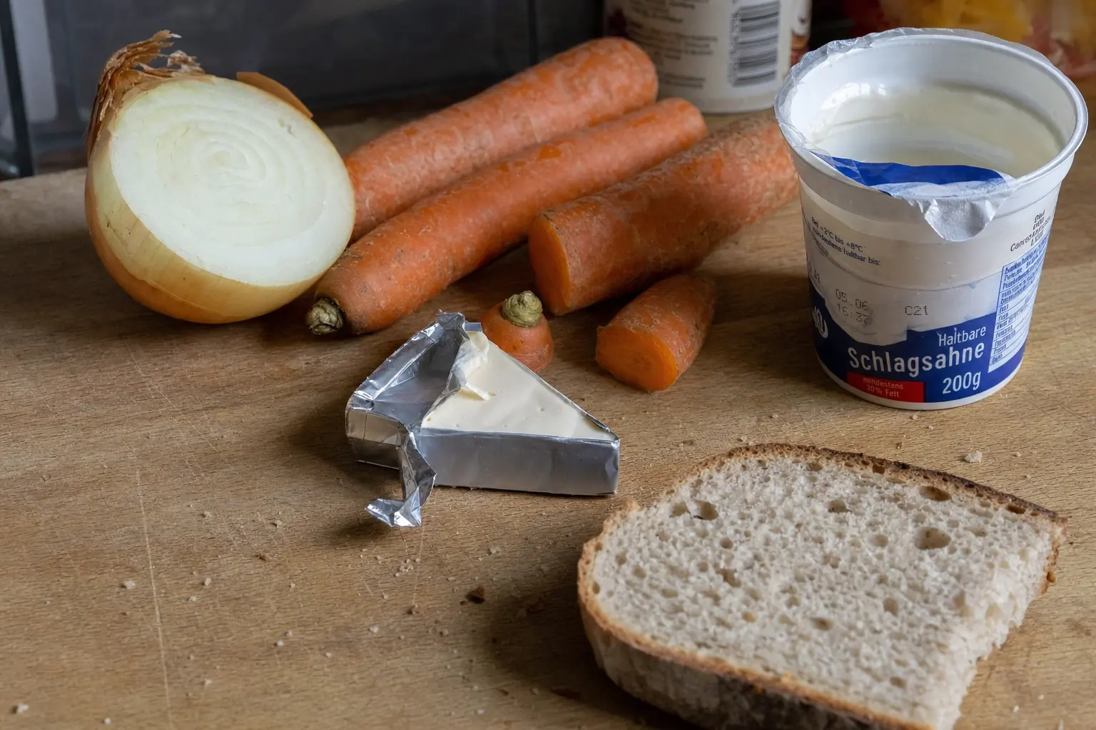 Leftovers on a kitchen table: half an onion, three carrots, a tub of heavy cream, a wedge of opened cheese, and two slices of bread